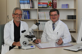 Dr. Jörg Gröticke (links), Leitender Oberarzt, und Prof. Dr. Bernd Hertenstein hinter einem Schreibtisch in Dienstkleidung mit Mikroskop