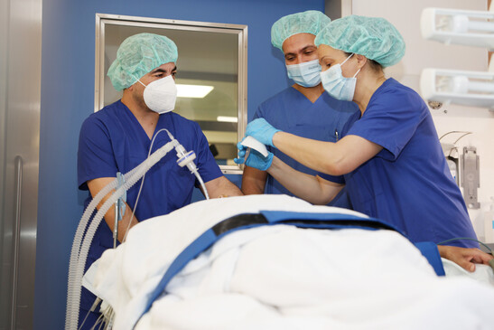 Drei medizinische Fachkräfte stehen in OP-Kleidung um ein Bett herum