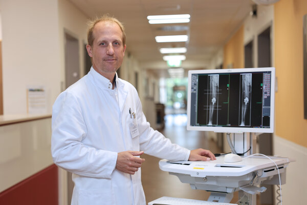 Dr. Knut Müller-Stahl in weißer Dienstkleidung mit Visitenwagen auf einem Stationsflur. Der Monitor auf dem Visitenwagen zeigt ein Bein als Röntgenbild