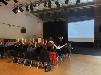 Das Veranstaltungsfoto zeigt im Vordergrund das Auditorium und im Hintergrund die Leinwand mit einer Vortragsfolie